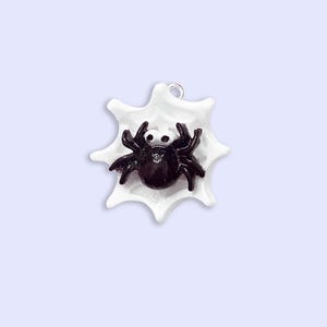 Op de afbeelding: Een kleine bedel met een glanzende zwarte spin met grote witte ogen, geplaatst in het midden van een wit, stervormig spinnenweb. Een klein zilverkleurig metalen lusje is aan de bovenrand van het witte web bevestigd om het op te hangen.