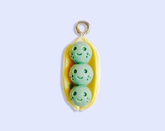 May include: A charm featuring three green peas with smiling faces inside a yellow pea pod. The charm has a silver loop for hanging.