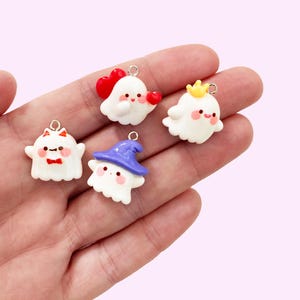 May include: Five white ghost-shaped charms with various accessories. One charm has a red bow tie, another a purple witch hat, and two hold red hearts. The last charm wears a gold crown. Each charm has a silver loop.
