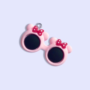 May include: Pink plastic sunglasses with red bows and black lenses. The sunglasses have a silver loop at the top for hanging.