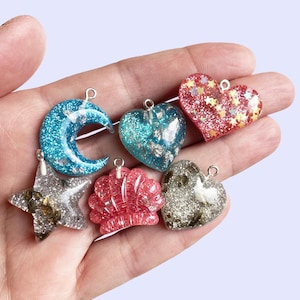May include: A collection of colorful resin charms in various shapes, including a crescent moon, hearts, a star, and a shell. The charms are filled with glitter and metallic accents, with a small loop for hanging.