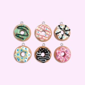 Könnte beinhalten: Sechs bunte Donut-Anhänger mit verschiedenen Belägen, darunter Streusel, Streifen und Schokoladen beträufelung. Die Anhänger sind aus Polymer Clay gefertigt und haben eine silberne Öse zum Aufhängen.