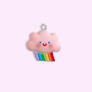 Puede incluir: Un dije en forma de nube rosa con una cara sonriente y un arcoíris. El dije tiene un lazo plateado en la parte superior para colgarlo. El arcoíris tiene rayas moradas, azules, verdes, amarillas y rojas.