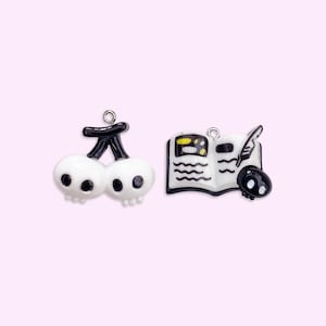 May include: Two charms with a spooky theme. One charm is a pair of white skulls shaped like cherries with a black stem. The other charm is a book with a quill and a skull. The charms are black and white.