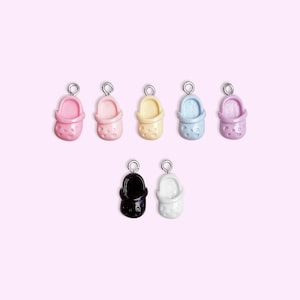 May include: Seven colorful miniature shoe charms in pink, peach, yellow, blue, and purple, plus black and white. Each charm has a silver-colored loop at the top. The charms are arranged on a light pink background.
