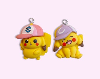 Puede incluir: Dos amuletos de Pikachu amarillos con sombreros de diferentes colores. Un amuleto tiene un sombrero rosa con un logotipo azul y blanco, y el otro amuleto tiene un sombrero morado con un logotipo blanco.