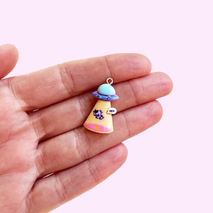 May include: A small UFO charm, held in a hand. The charm features a light blue top, a purple and blue rim, and a tan base. A cartoon cow is on the base, with a speech bubble that says "MOO".