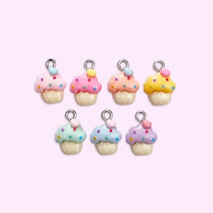 Op de afbeelding: Zeven kleurrijke cupcake-bedels met zilverkleurige lussen voor het maken van sieraden. Elke cupcake heeft een crèmekleurige basis en een andere pastelkleurige frosting, versierd met kleine, kleurrijke hagelslag en een kers.