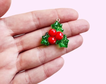 May include: A Christmas-themed charm featuring a holly sprig with three bright red berries and green leaves. The charm has a gold-colored loop for attaching to jewelry or other crafts. The design is set against a soft pink background.