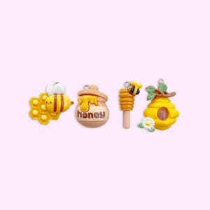 May include: Four small, colorful, clay charms with a bee theme. The charms include a bee on a honeycomb, a honey pot, a honey dipper, and a beehive.