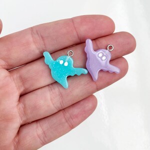 May include: Two ghost-shaped charms featuring a smooth, glittery finish. One charm is light blue and the other is light purple. Each ghost has two small white circular eyes and a silver-toned metal jump ring attached to the top for hanging.