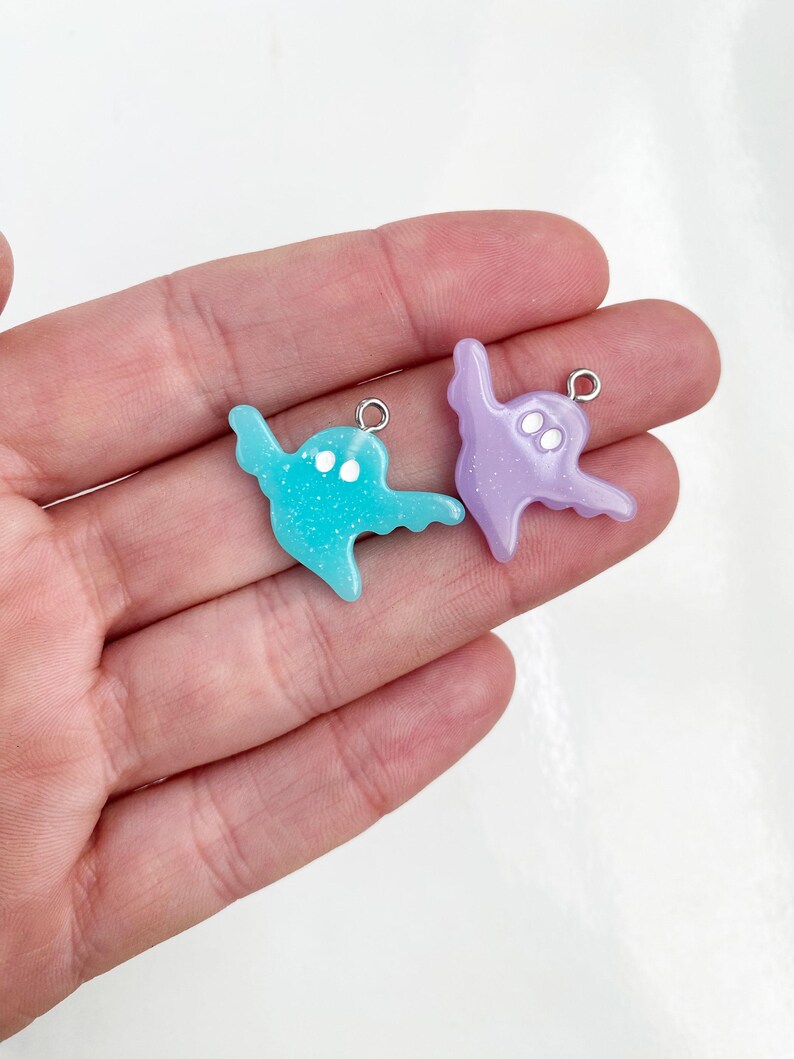 May include: Two ghost-shaped charms featuring a smooth, glittery finish. One charm is light blue and the other is light purple. Each ghost has two small white circular eyes and a silver-toned metal jump ring attached to the top for hanging.