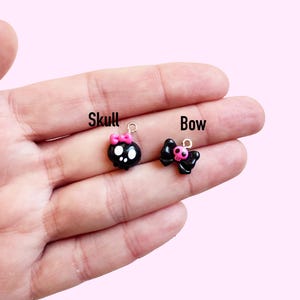 May include: Two small, black charms are displayed. One is a skull with a pink bow, and the other is a bow with a pink center. The word "Skull" is written above the skull charm, and the word "Bow" is written above the bow charm.