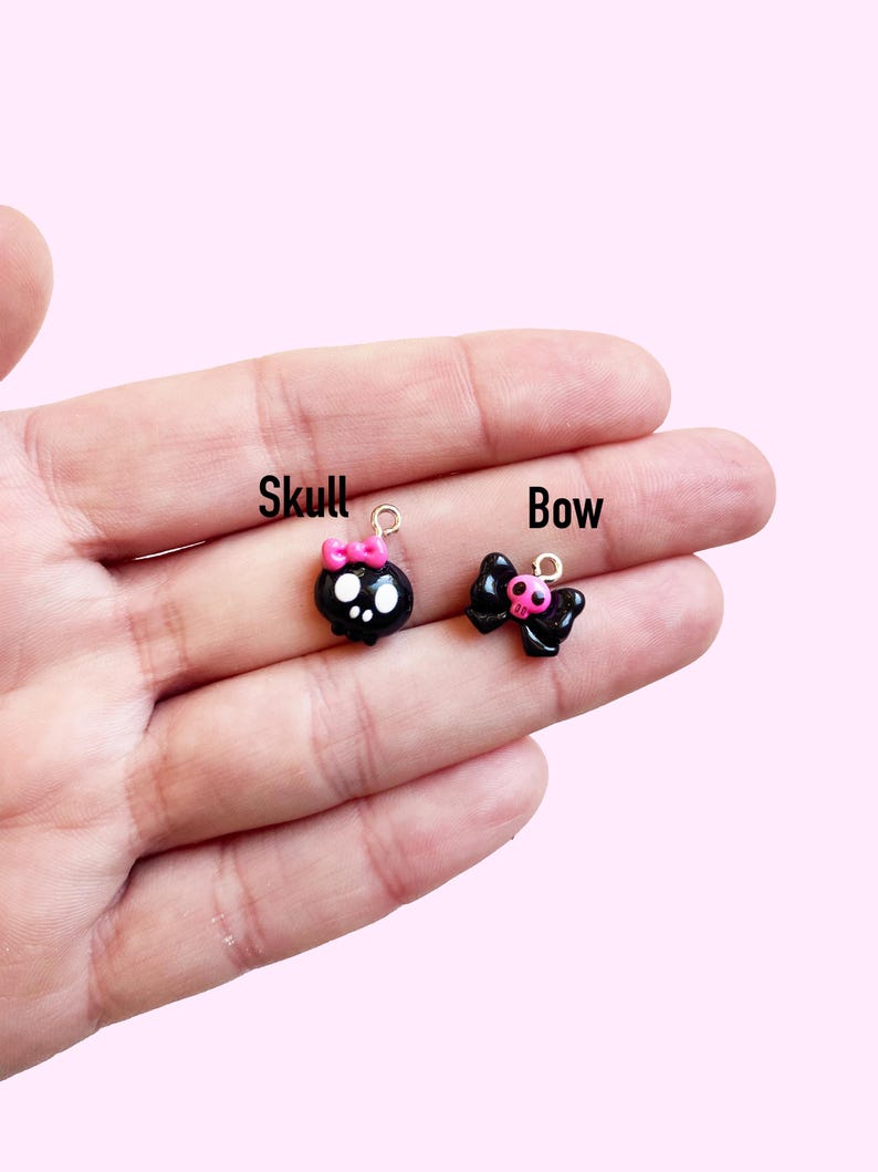 May include: Two small, black charms are displayed. One is a skull with a pink bow, and the other is a bow with a pink center. The word "Skull" is written above the skull charm, and the word "Bow" is written above the bow charm.