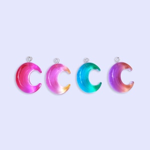 May include: Four colorful crescent moon charms in shades of pink, orange, blue, and purple. Each charm has a silver loop at the top for hanging. The background is a light purple.