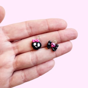 May include: Two small black skull charms with pink accents. One skull has a pink bow, the other has a pink bow on top. Both charms have gold-colored loops for attaching to jewelry or other crafts.