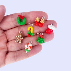 May include: A collection of seven small Christmas-themed charms including a green wreath with a red bow, reindeer antlers with a red bow, a red stocking with white trim, a gingerbread person, a candy cane with holly, a green tree, and a lollipop.