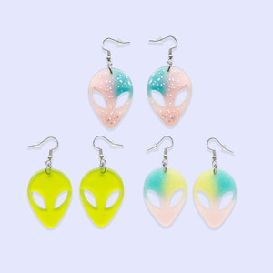 May include: A set of six alien head-shaped earrings. Two pairs are pink and blue with glitter, and two pairs are solid yellow.