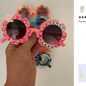 May include: Pink sunglasses with round frames decorated with iridescent pink beads and a white ghost charm. The frames feature white letter beads spelling GHOUL GANG around the lenses. Additional pairs in orange, blue, and black are visible behind.