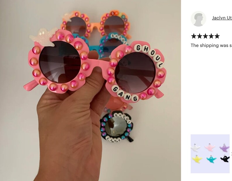 May include: Pink sunglasses with round frames decorated with iridescent pink beads and a white ghost charm. The frames feature white letter beads spelling GHOUL GANG around the lenses. Additional pairs in orange, blue, and black are visible behind.