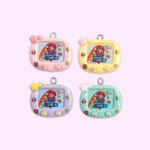 May include: Four colorful Super Mario themed pendants. Each pendant features a small screen with Mario, stars, and the words "Super Mario". The pendants are in pink, yellow, purple, and mint green, with a silver loop for hanging.