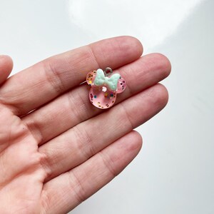 May include: A small, translucent pink resin charm shaped like a mouse head with rounded ears. The charm is filled with multicolored glitter flakes and features a pale mint green bow attached to the top, complete with a small silver metal jump ring.