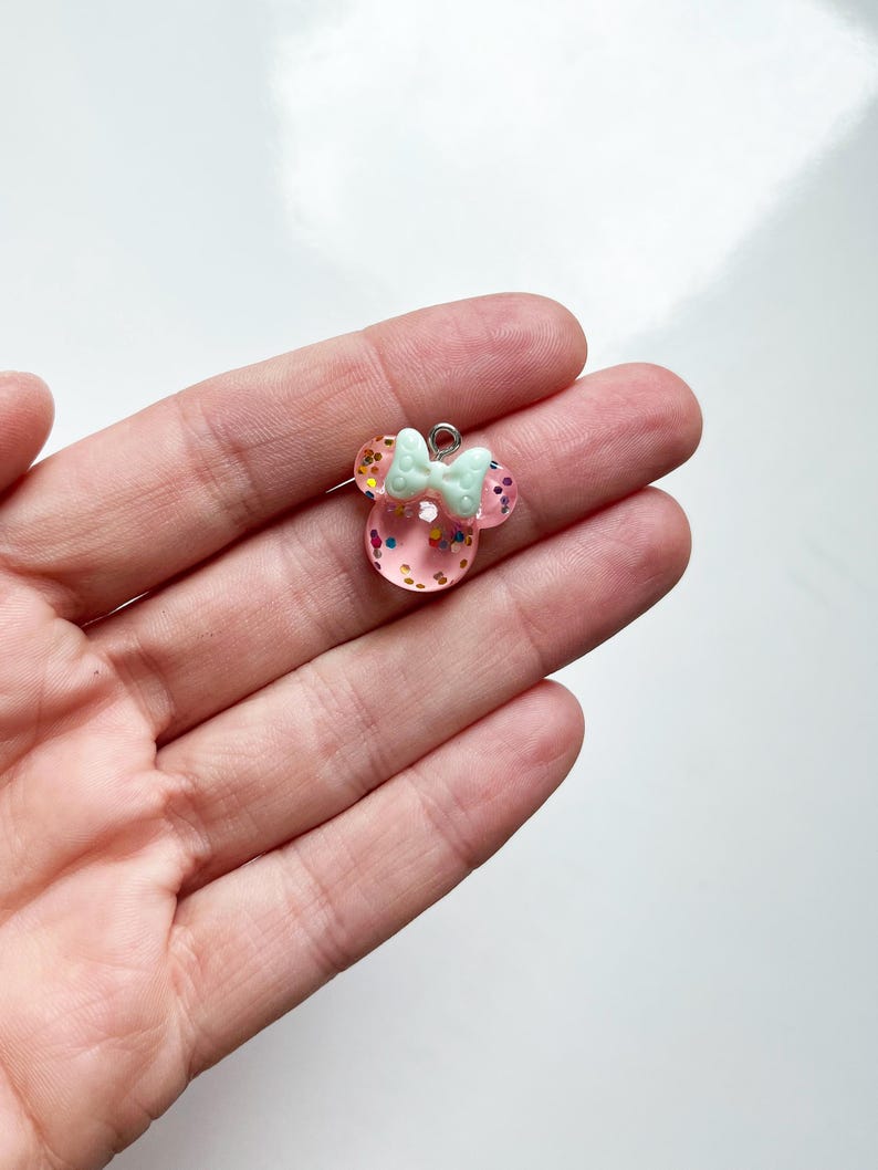May include: A small, translucent pink resin charm shaped like a mouse head with rounded ears. The charm is filled with multicolored glitter flakes and features a pale mint green bow attached to the top, complete with a small silver metal jump ring.