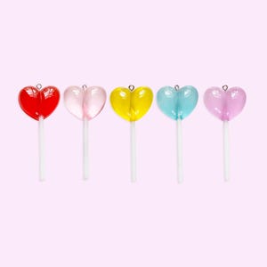 May include: Five heart-shaped lollipops in red, pink, yellow, blue, and purple, each with a white stick. The lollipops are arranged in a row against a light pink background. These are perfect for Valentine's Day.