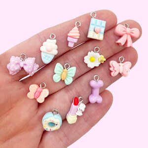 May include: A collection of 12 pastel-colored charm pendants featuring various cute designs, including a butterfly, a cupcake, a donut, a lipstick, a bone, a bow, a flower, and a chocolate bar.