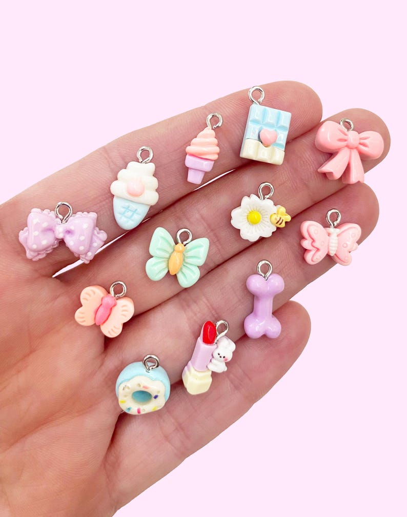 May include: A collection of 12 pastel-colored charm pendants featuring various cute designs, including a butterfly, a cupcake, a donut, a lipstick, a bone, a bow, a flower, and a chocolate bar.
