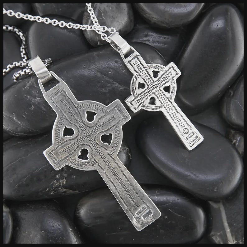 May include: Two silver Celtic cross pendants on chains. The crosses have detailed knotwork designs and are set against smooth, dark stones. The pendants feature cut-out shapes and textured surfaces, reflecting traditional Celtic artistry.