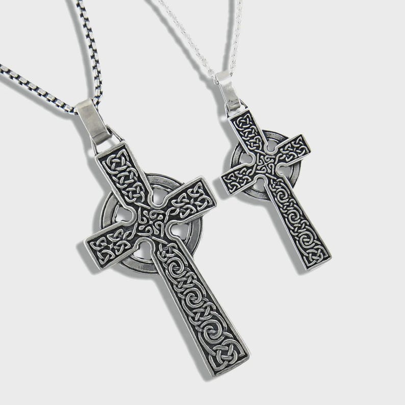 May include: Two silver Celtic cross pendants featuring detailed knotwork patterns. Each cross hangs from a chain, one with a black and silver chain and the other with a silver chain. The crosses have a circular element at the centre.