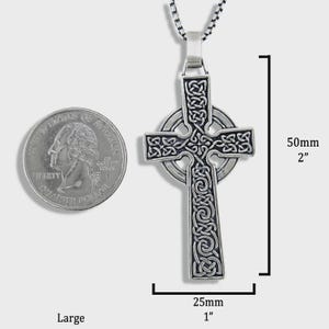 May include: A silver Celtic cross pendant featuring detailed knotwork. The pendant measures 50mm in length and 25mm in width. The cross hangs from a silver chain. A quarter coin is shown for scale.