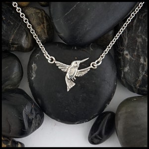 Puede incluir: Un collar de plata con forma de colibrí con una cadena delicada. El colibrí está en vuelo con las alas extendidas. El collar se muestra sobre un lecho de piedras negras.