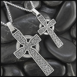 May include: Two silver Celtic cross pendants with detailed knotwork patterns, each hanging from a fine chain. The crosses are displayed on a bed of smooth, dark grey stones, providing a strong contrast. The pendants have an intricate, openwork design.