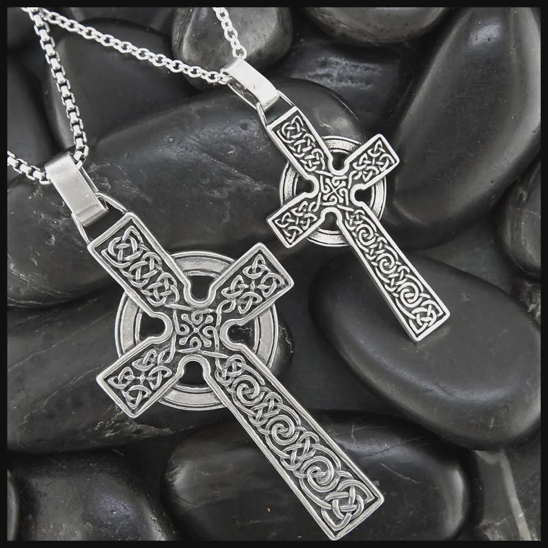 May include: Two silver Celtic cross pendants with detailed knotwork patterns, each hanging from a fine chain. The crosses are displayed on a bed of smooth, dark grey stones, providing a strong contrast. The pendants have an intricate, openwork design.