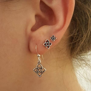 Puede incluir: Primer plano de una oreja adornada con tres pendientes de plata. Uno es un pendiente colgante con un diseño de nudo celta, y dos son pendientes de botón con un diseño de nudo similar. Los pendientes tienen un centro rojo.