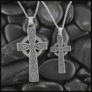 May include: Two silver Celtic cross pendants on chain necklaces. The crosses have intricate knotwork designs, set against dark, smooth stones. The pendants are different sizes, showcasing detailed craftsmanship.
