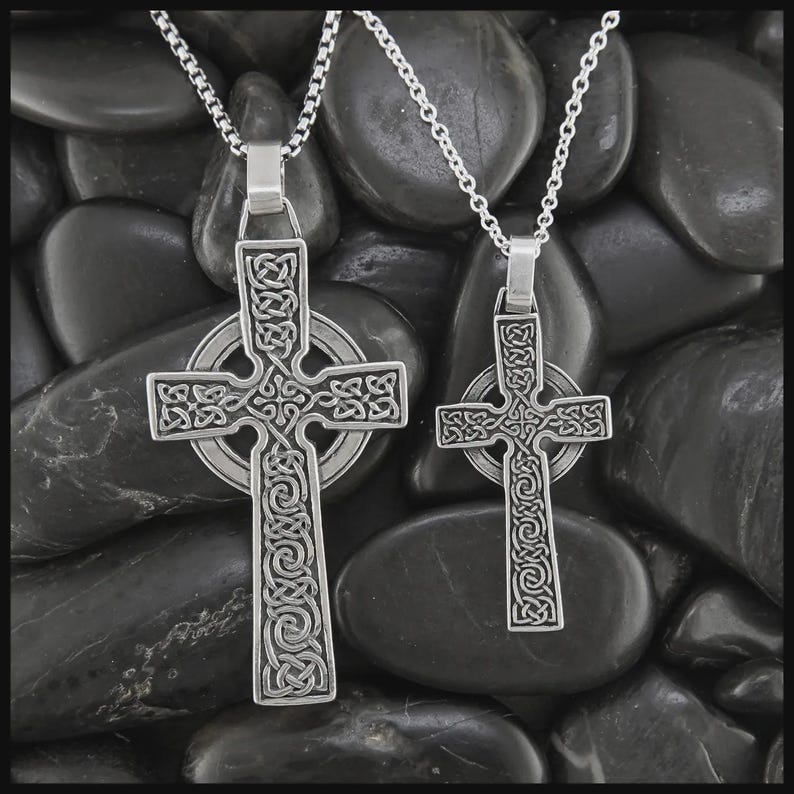 May include: Two silver Celtic cross pendants on chain necklaces. The crosses have intricate knotwork designs, set against dark, smooth stones. The pendants are different sizes, showcasing detailed craftsmanship.