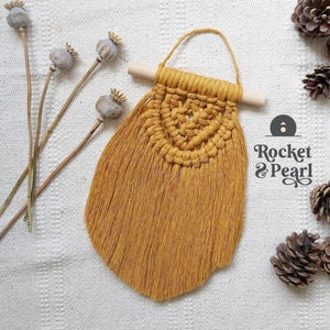 Puede incluir: Un tapiz de macramé de color amarillo mostaza con flecos. El tapiz está sujeto a una varilla de madera con un lazo para colgar.