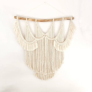 May include: A white macrame wall hanging with a wooden dowel at the top and a fringe design. The hanging is made of cotton cord and has a bohemian style.