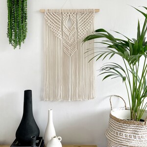 May include: A white macrame wall hanging with a geometric design, hanging on a white wall. There are three vases on a wooden table in front of the wall hanging, one black, one white, and one cream. A potted plant in a woven basket is on the right side of the image.