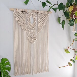 May include: A macrame wall hanging with a geometric design in a cream color. The hanging is made with natural cotton cord and has a wooden dowel at the top.
