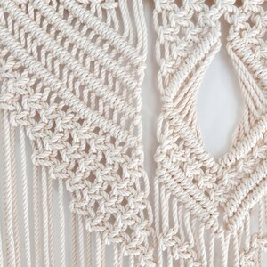 May include: Close-up of a macrame wall hanging with a white cotton cord. The hanging features a geometric pattern with a large open space in the center.