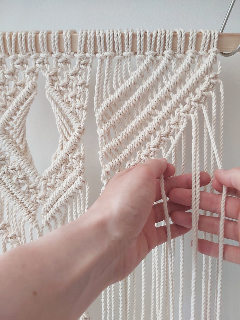 May include: A close-up of a white macrame wall hanging with a geometric pattern. The macrame is being held by a hand, showing the intricate knotting.