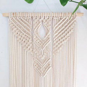 May include: A macrame wall hanging with a geometric design in a cream color. The hanging is made with natural cotton cord and is attached to a wooden dowel.