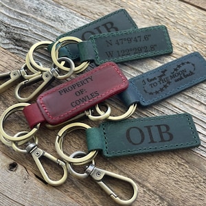 May include: Four leather keychains with different engravings. One keychain is red and says "PROPERTY OF: COWLES". Another keychain is blue and says "I love you to the moon and back". A third keychain is green and says "OIB". The fourth keychain is green and says "N 47°9'47.6" W 122°29'2.8"".