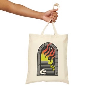 May include: Cream-colored tote bag featuring a graphic of a stone archway with stairs, flames, and a skull. The flames are red, orange, and yellow. The tote bag is held by a person.