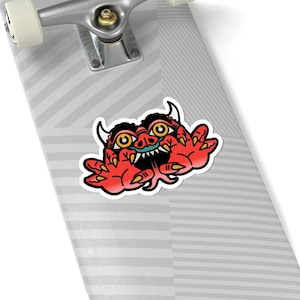 May include: A red sticker featuring a cartoon demon with yellow eyes, white horns, and a green mouth. The sticker has a black outline and is placed on a skateboard.