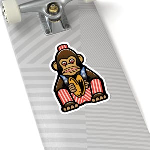 May include: A sticker featuring a cartoon monkey with a patriotic theme. The monkey is brown with a red and white striped hat and legs, and a vest with blue stars. It holds a yellow musical instrument.